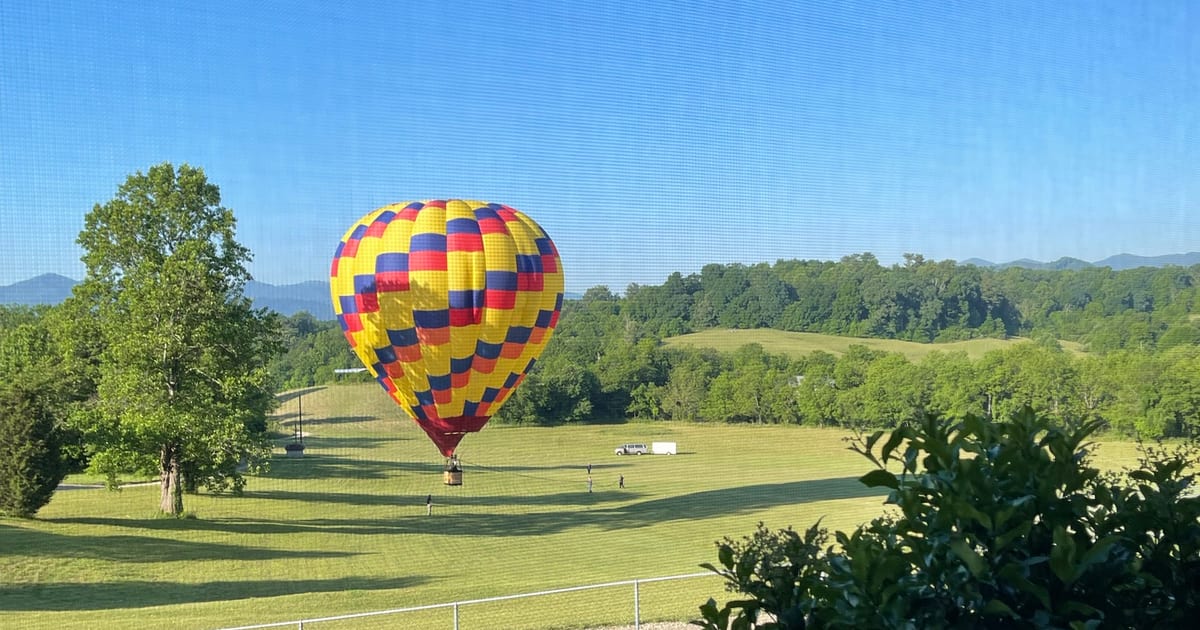 A red, yellow and blue hot air balloon landing in a green field in Candler, NC, with the Blue Ridge Mountains behind it