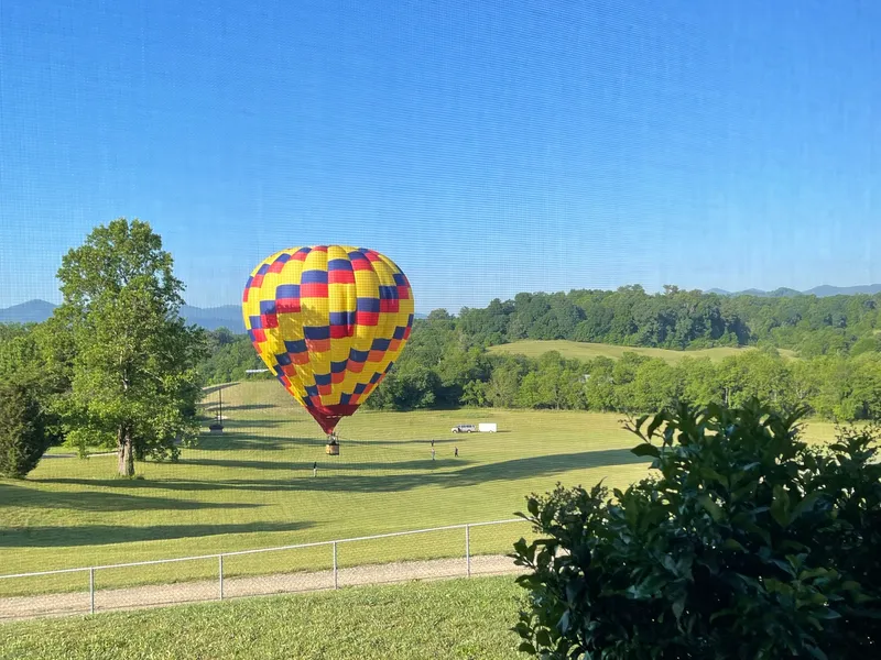 A hot air balloon touching down in a Candler field on a summer morning, the chase crew walking out to it, mountains behind